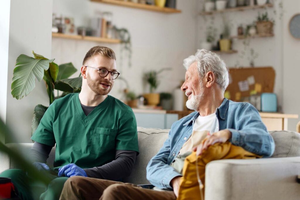 An Amerita caregiver sits with an older patient on a couch at home